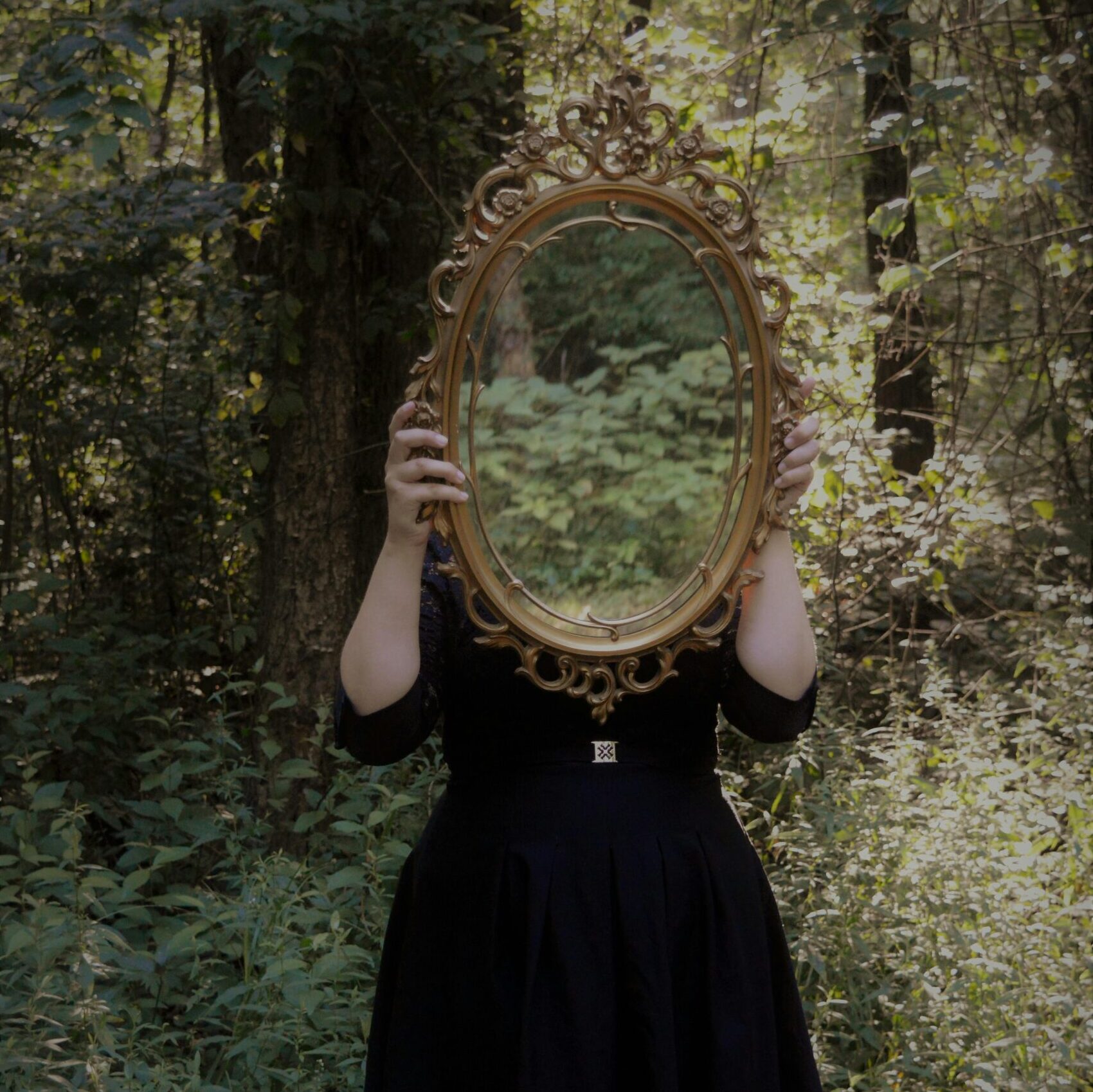 A woman holds an ornate mirror in a forest setting, reflecting nature.